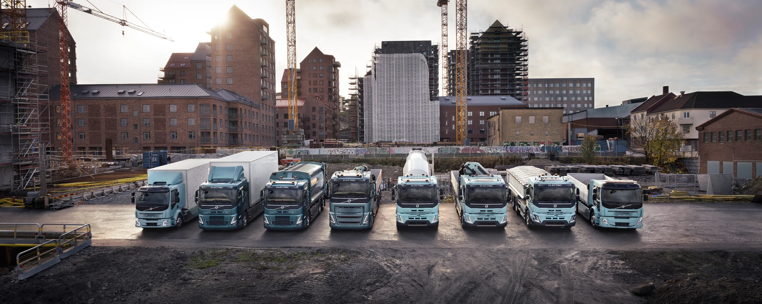 Åtte ulike elektriske lastebiler står på rekke, oppstilt foran heisekran og urbant miljø