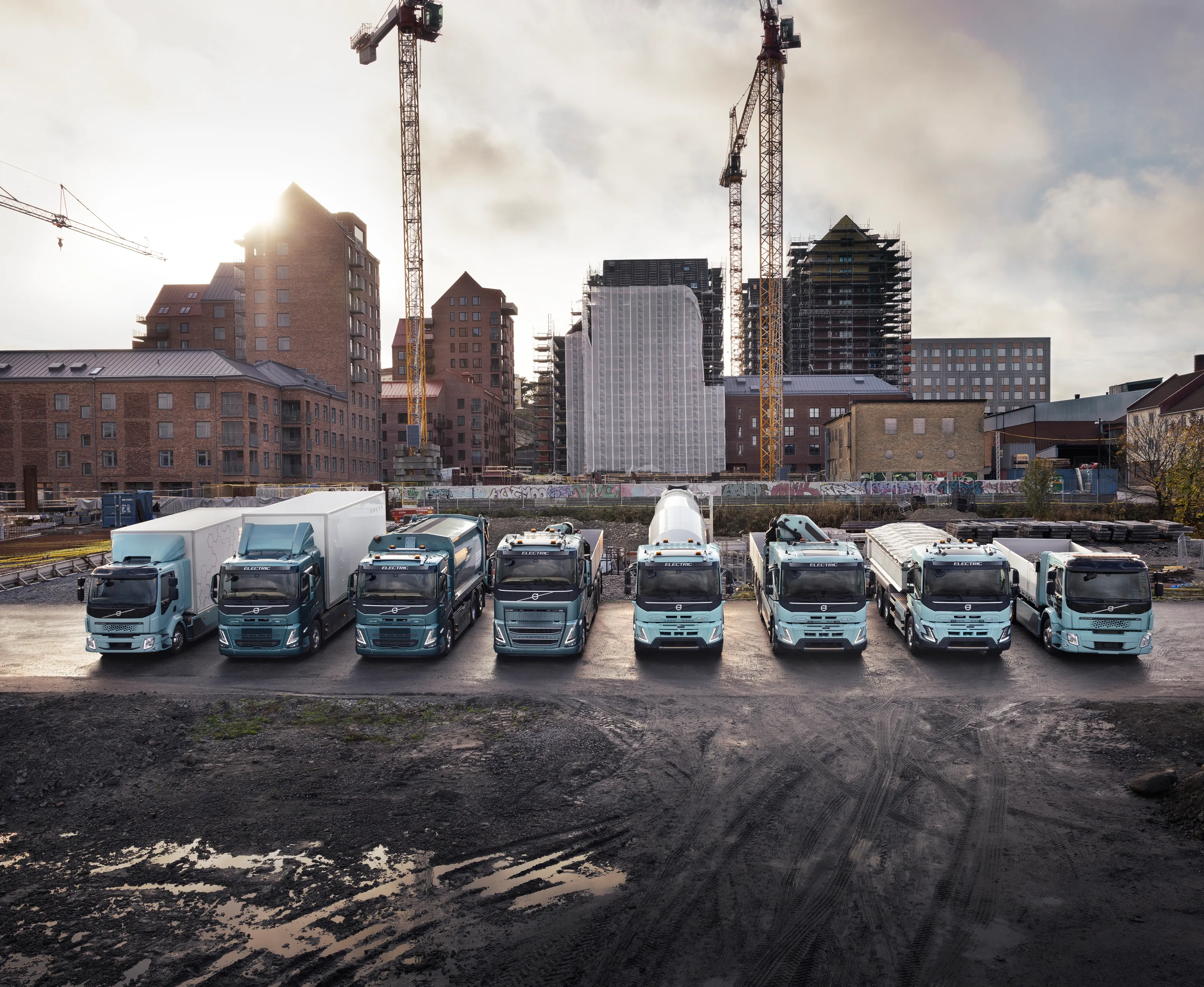 Åtte ulike elektriske lastebiler står på rekke, oppstilt foran heisekran og urbant miljø