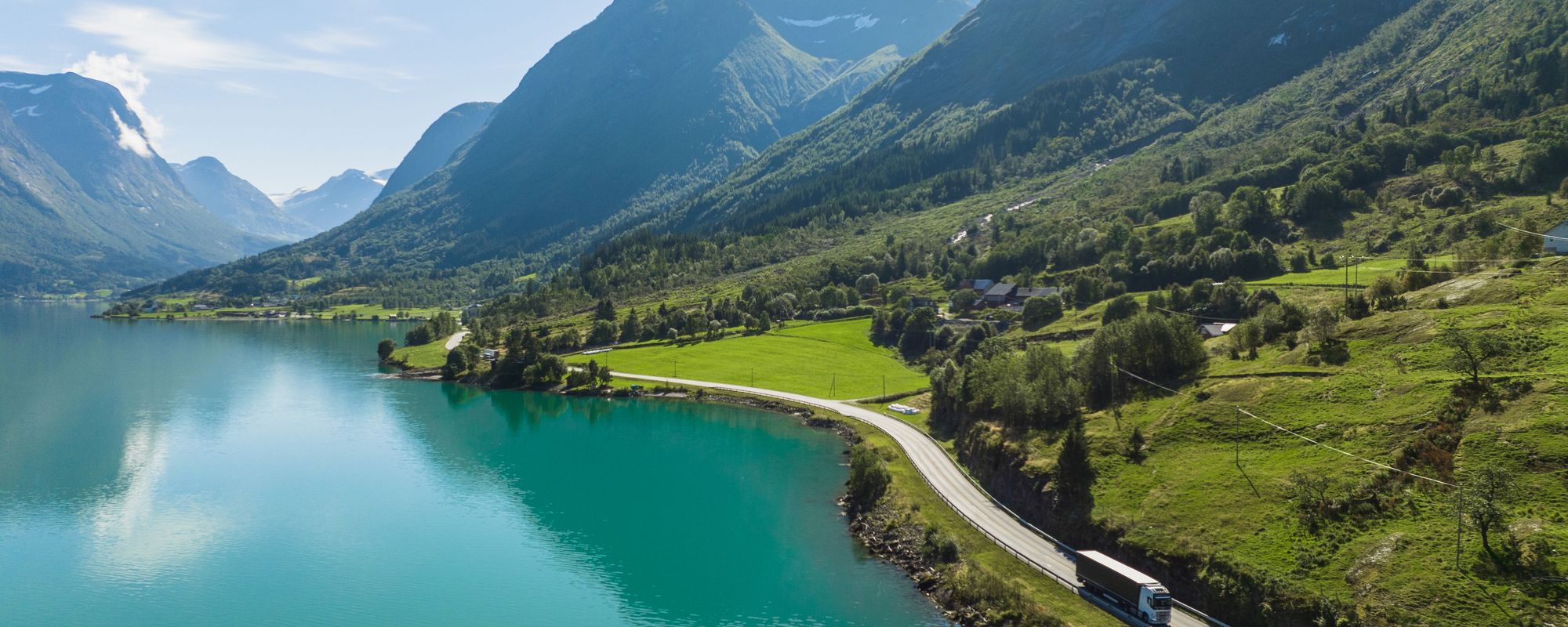 En lastebil kjører langs en idyllisk norsk fjord, med bratte fjell rundt