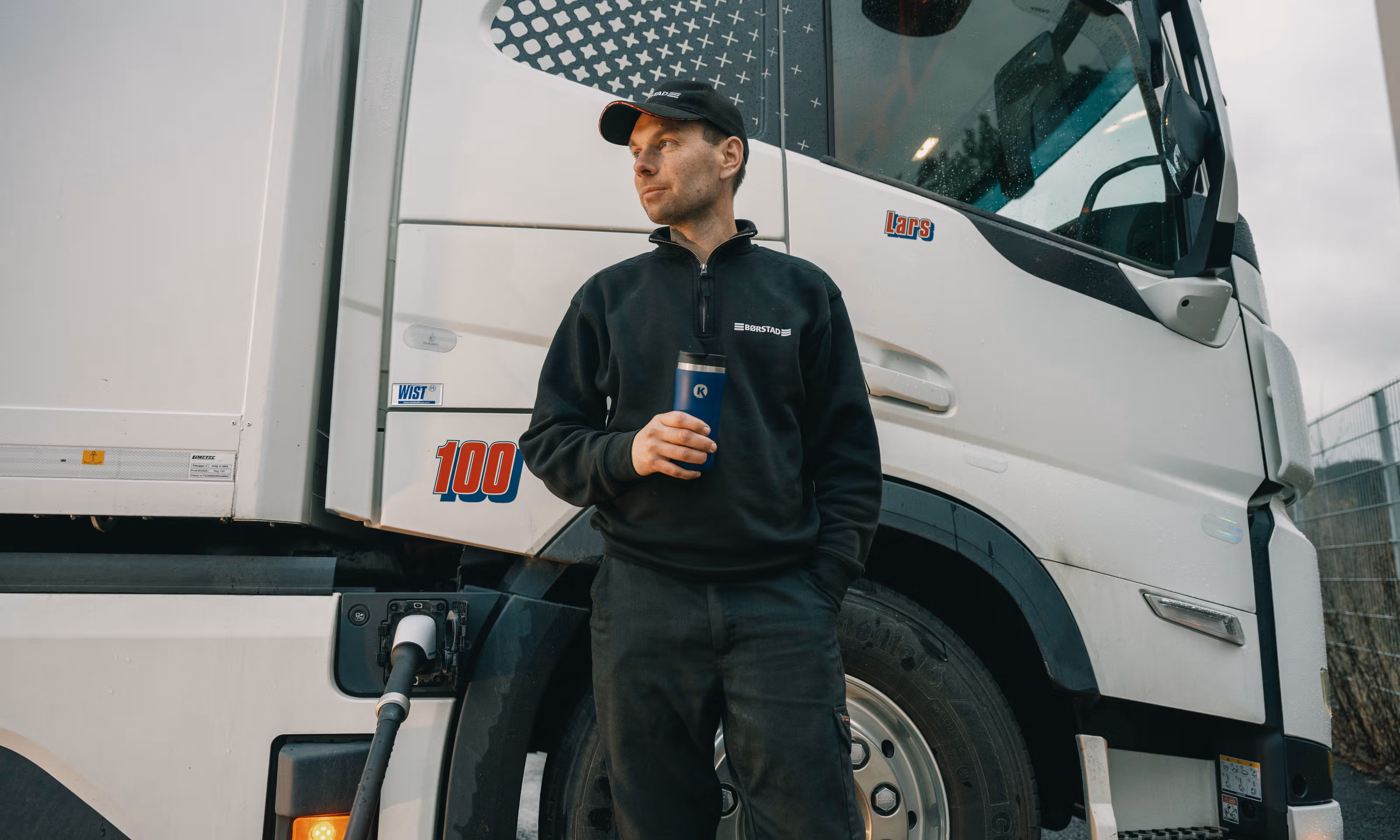 Mann i caps drikker kaffe mens han lader sin elektriske lastebil.