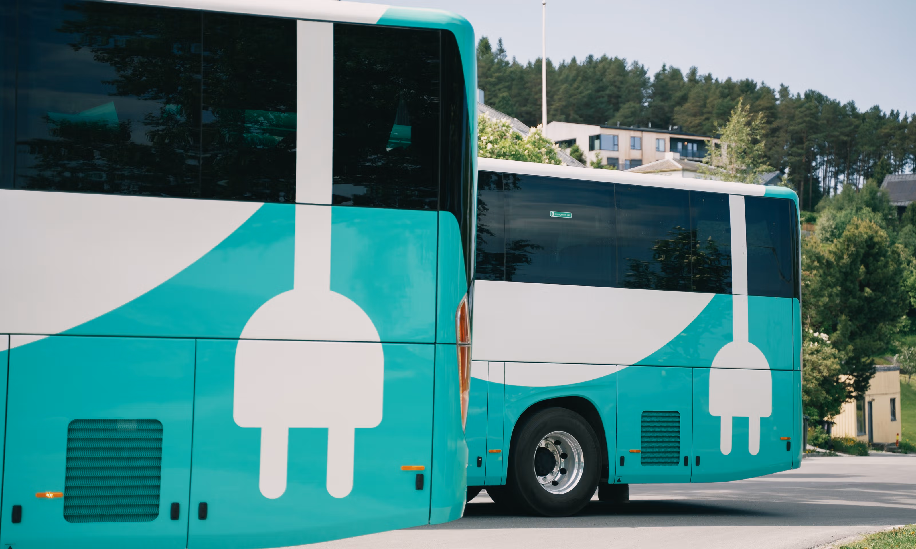 To oppstilte elektriske busser som det vises kun bakre del av, med enebolig og skog i bakgrunnen.