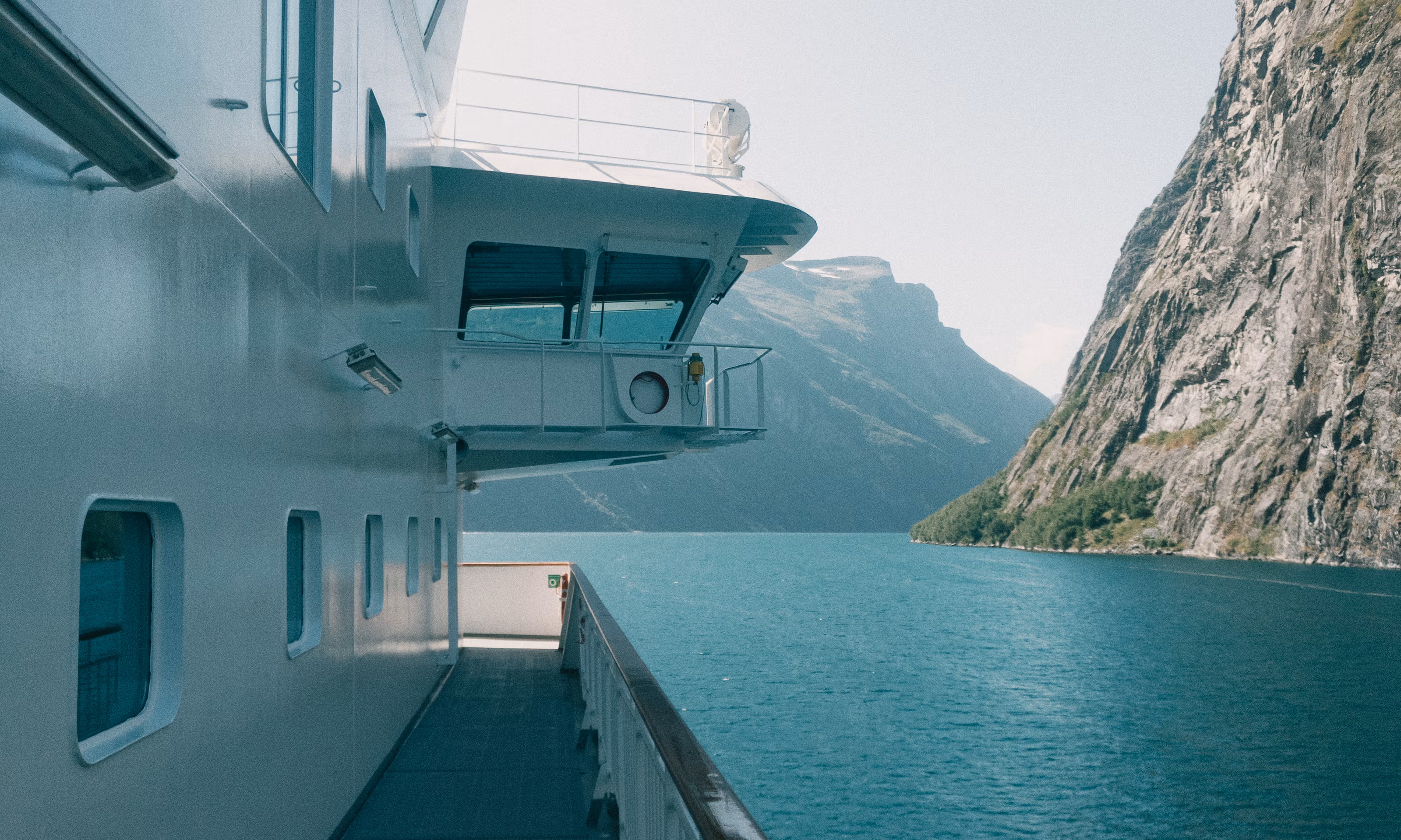 En passasjerbåt sett fra siden, inn i en fjordarm med asurblå sjø og en fjellvegg på høyre side.