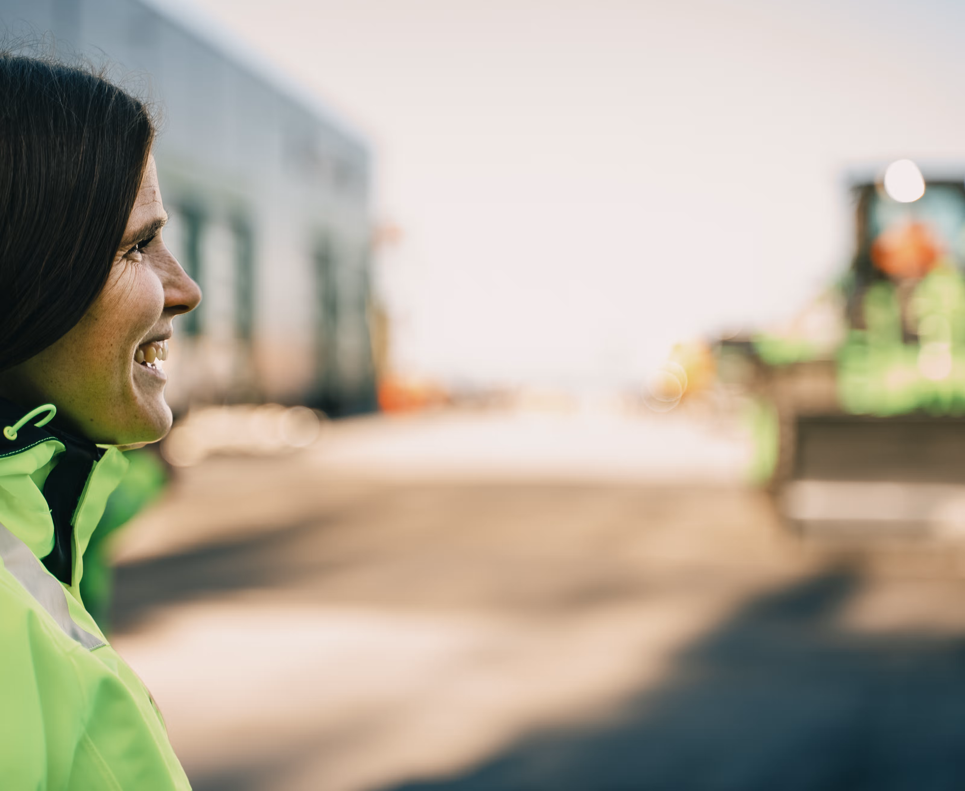 En kvinne i arbeidstøy som smiler, med en uskarp anleggsmaskin i bakgrunnen.