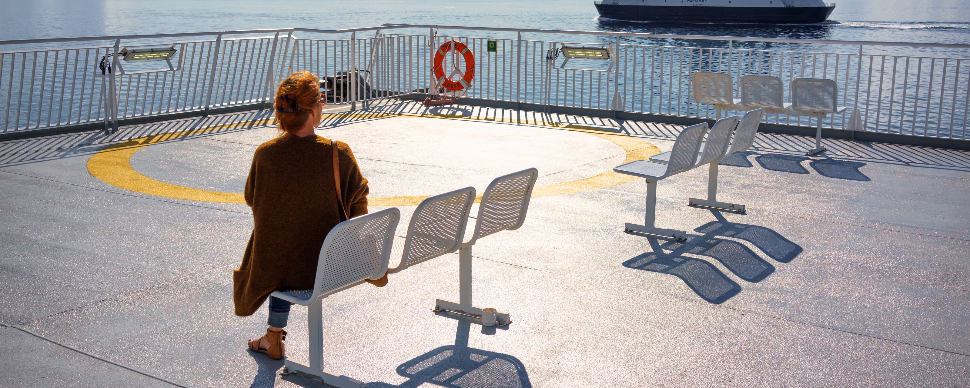 En kvinne sitter på dekk i sollyset og ser på en annen ferje kjører forbi.