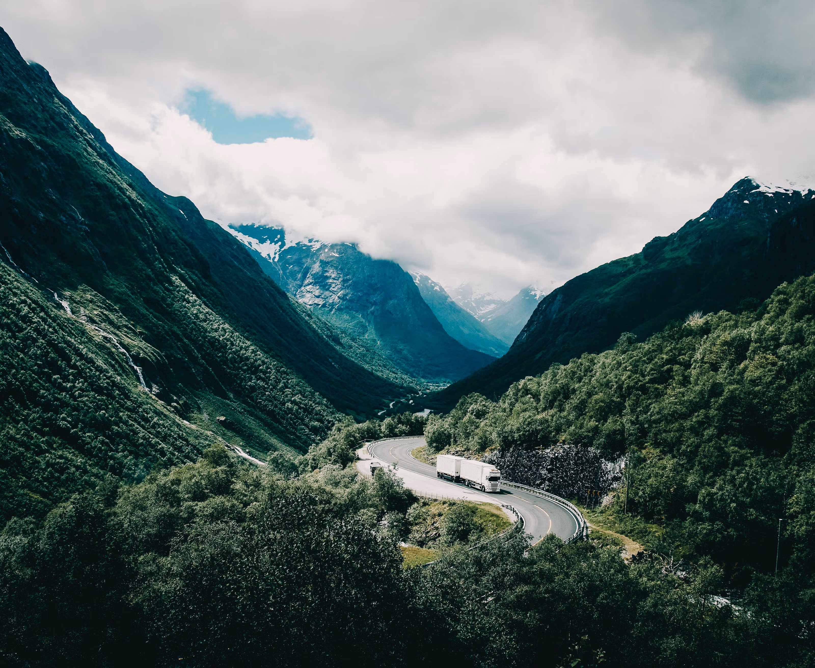 En lastebil som svinger seg igjennom et storslått, grønt, norsk landskap med fjell i bakgrunnen.