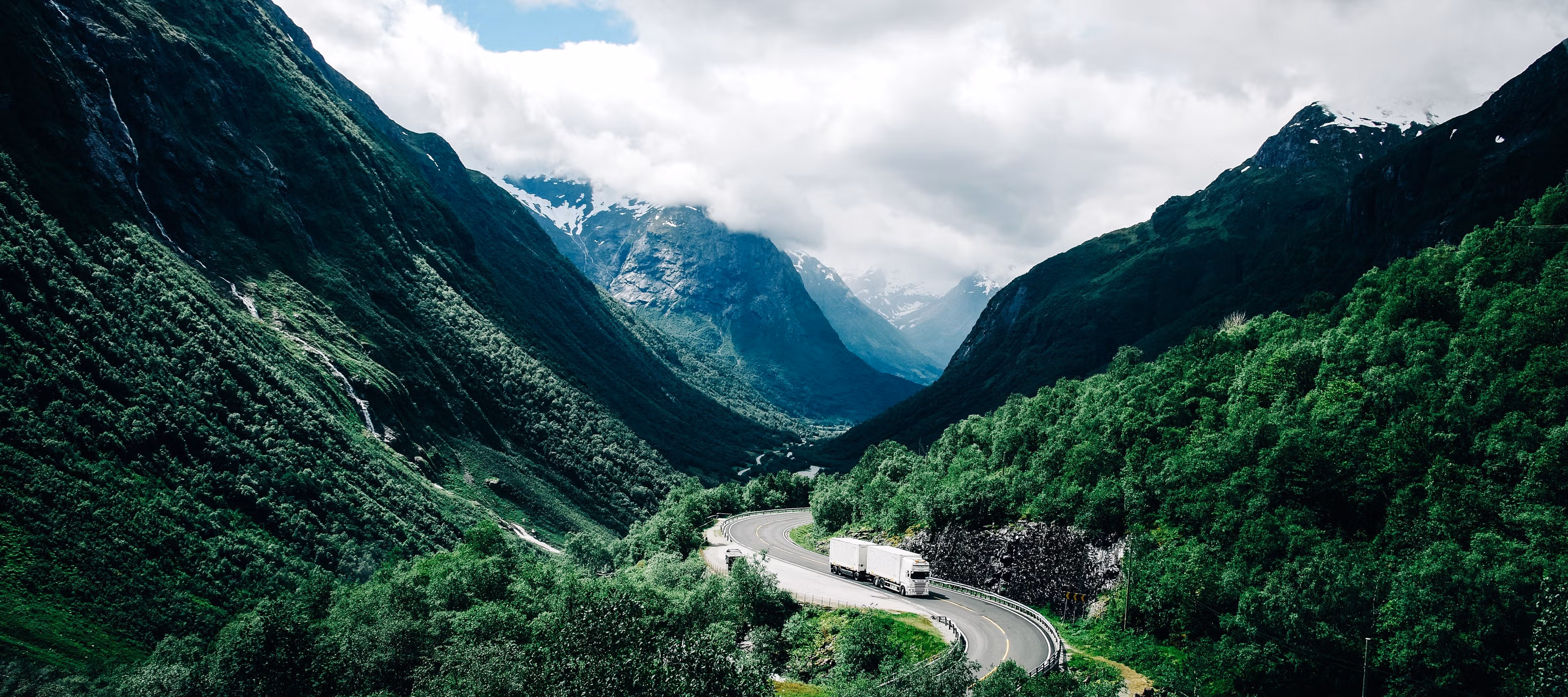 En lastebil som svinger seg igjennom et storslått, grønt, norsk landskap med fjell i bakgrunnen.
