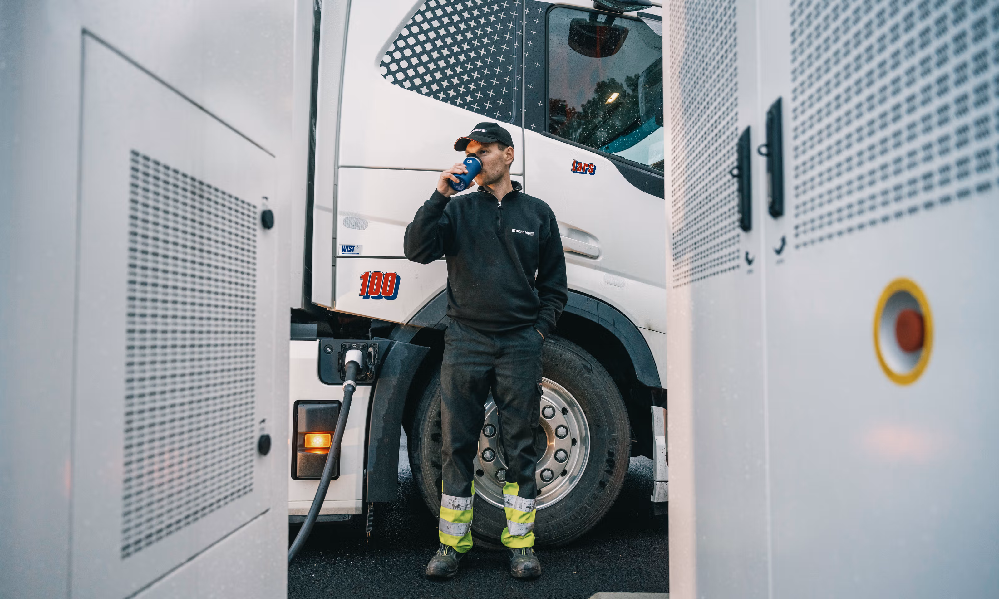 Mann i caps drikker kaffe mens han lader sin elektriske lastebil.
