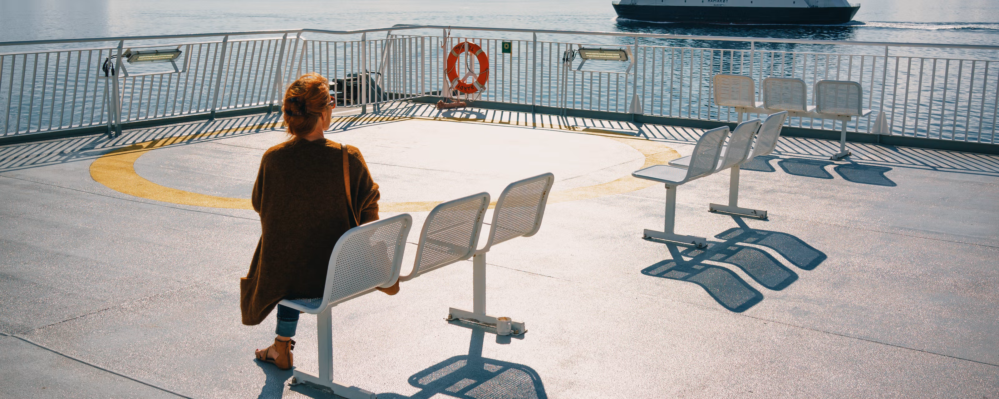 En kvinne sitter på dekk og ser en annen ferge ut på fjorden.